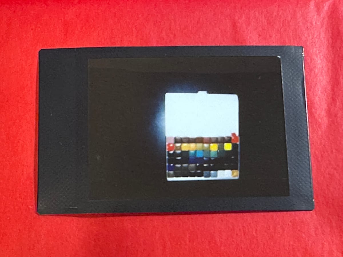 A black framed Instax photo on a red background. The subject of the Instax is an open palette of a LOT of small pans of watercolour on a dark backround, it's offset in the Instax image.