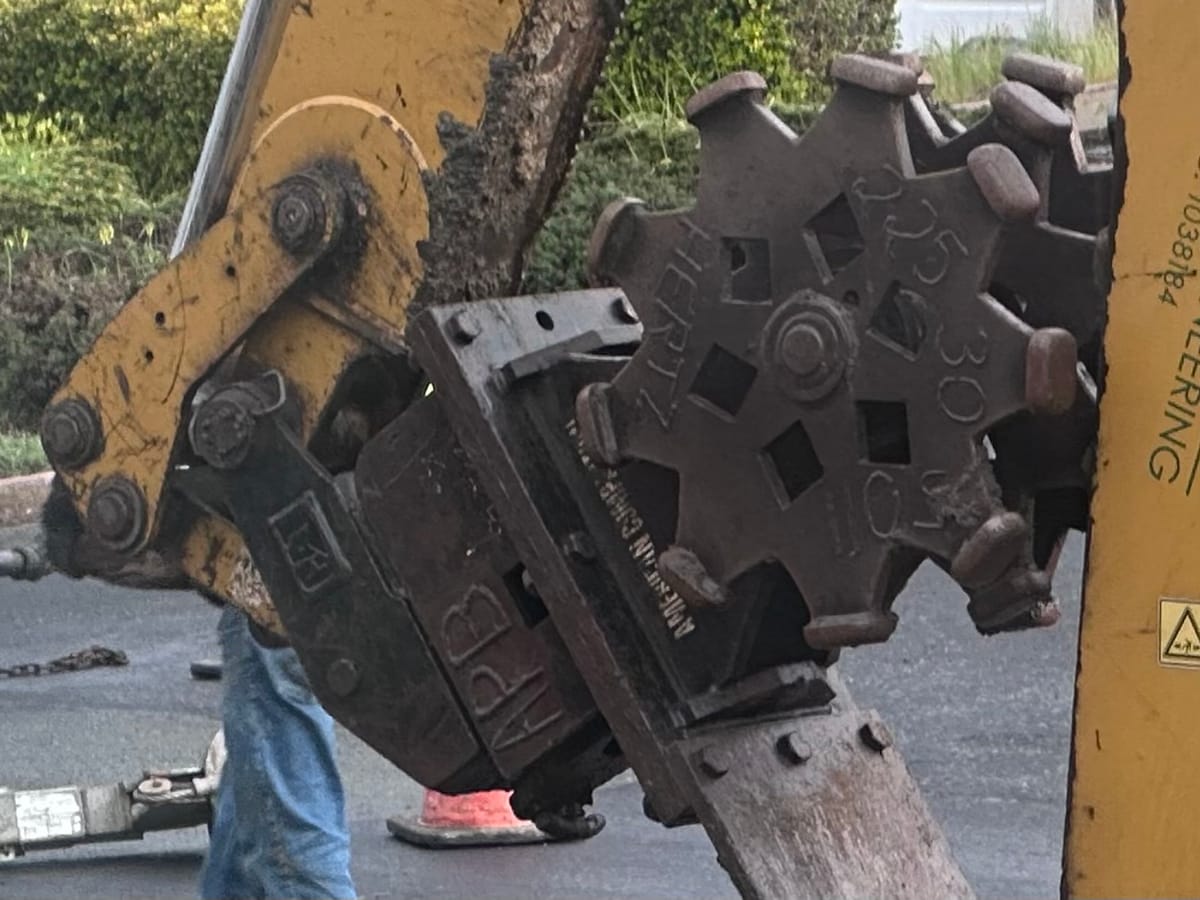 Some sort of huge grinding digger wheels on a hydraulic arm with debris in the background.