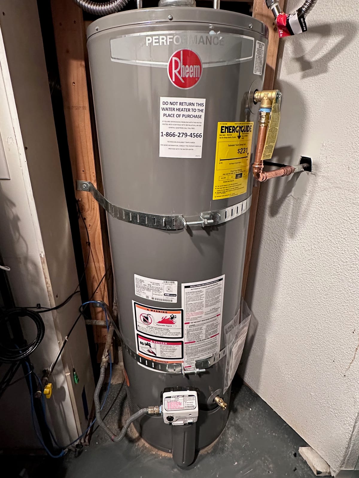 A new shiny grey tank water heater with pipes, clamps, hoses all with tags and new stickers all over it, set into a basement corner. It says "performance rheem" in a shiny label at top.