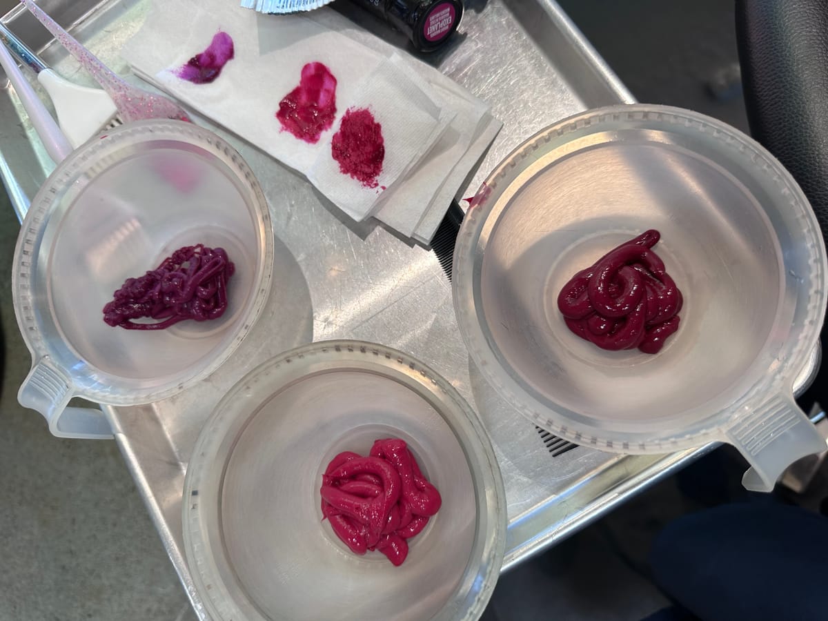 Three hairdressing bowls filled with various shades of pink hair dye on a metal stand. There's other various hair dye tools & a paper towel with two pink & one purple colour swatched out.