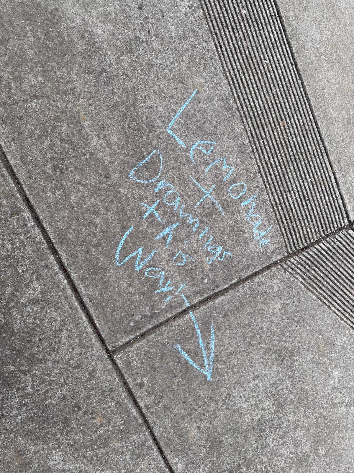 Portrait photo of a sidewalk with blue chalk on it that says "Lemonade + Drawings this way!" with an arrow pointing down.
