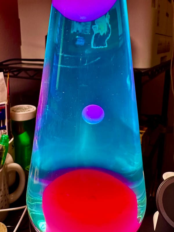 Closeup of a lava lamp with blue fluid and pink lava in mid bloop. You can see some desk cruft in the background around it. A laser printer on a stand is visible behind the fluid.