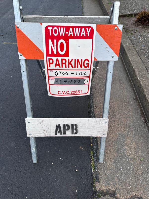 an a-frame in a wet street. APB is stencilled on the bottom crossbar & the top has a "tow away No Parking" sign. The time is 0700 - 1700 the date is duct-tape w/sharpie 2-17-26 to 3-27-26