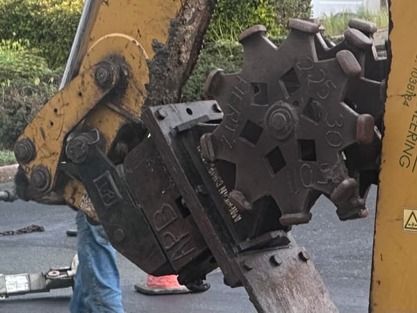 Some sort of huge grinding digger wheels on a hydraulic arm with debris in the background.
