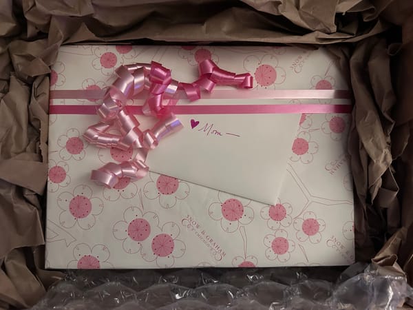 A wrapped box in white paper with pink and fuchsia flowers printed on it, curly ribbon, a card with handwritten "mom" on it, all nestled in packing paper.