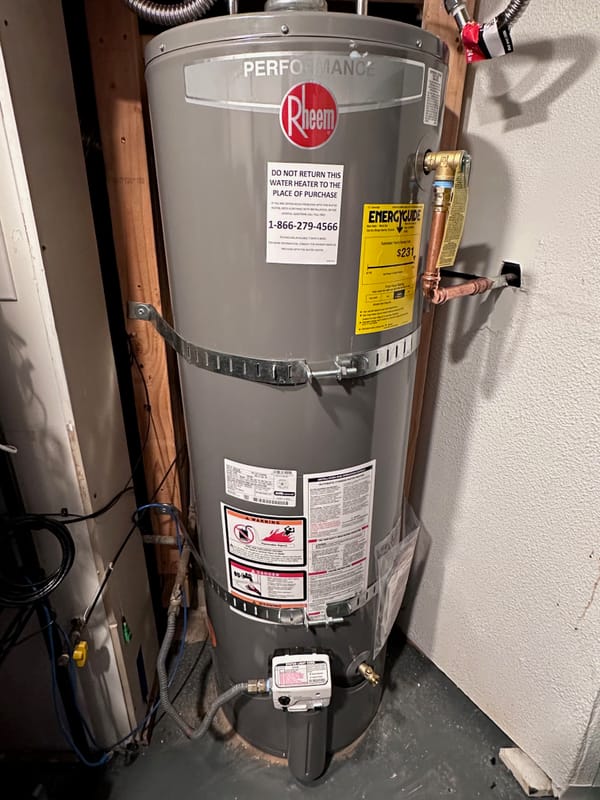 A new shiny grey tank water heater with pipes, clamps, hoses all with tags and new stickers all over it, set into a basement corner. It says "performance rheem" in a shiny label at top.